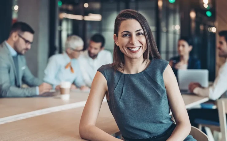 A smiling person sitting at a conference table with other people in the background.