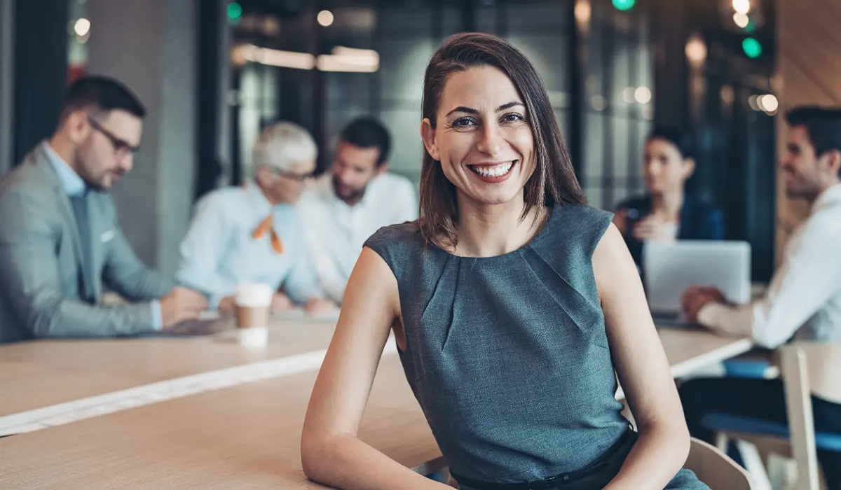 A smiling person sitting at a conference table with other people in the background.