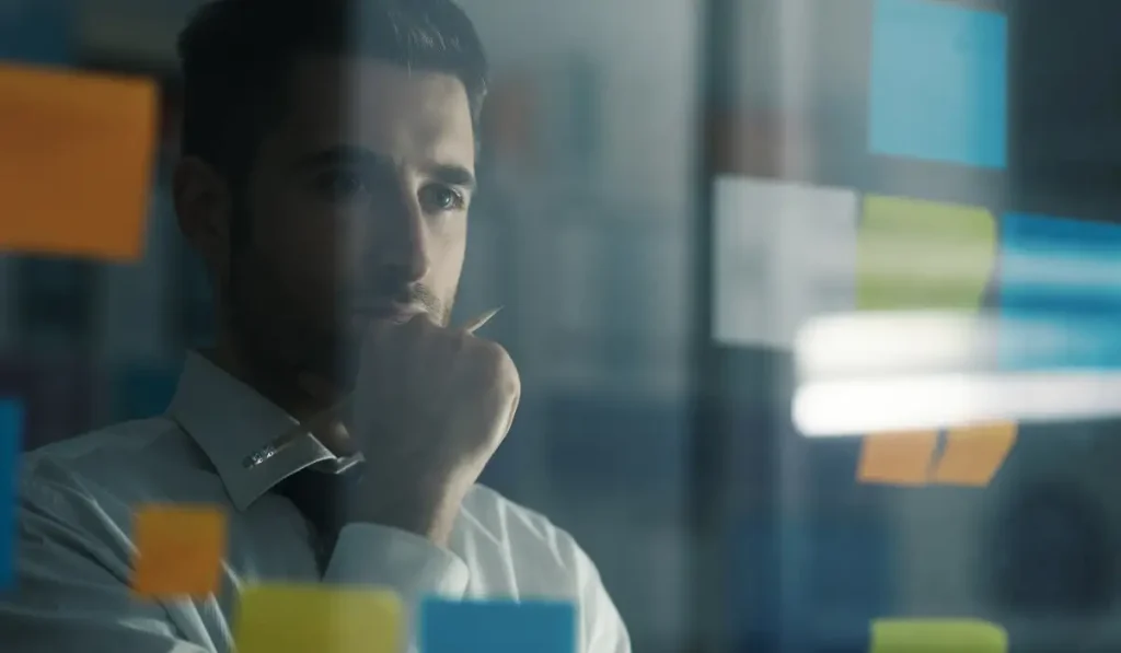 a person thinking while looking at sticky notes placed on a glass wall.